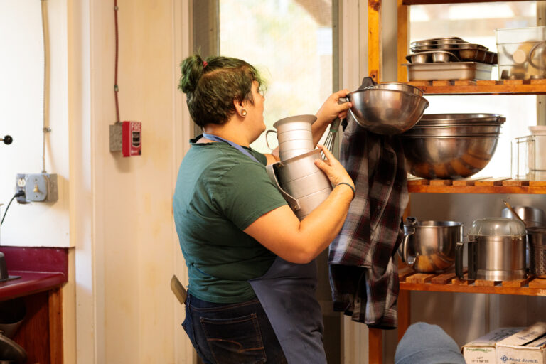 A person wearing a kitchen apron with a spatula in their back jeans' pocket grabs mixing bowls and measuring cups from kitchen shelves